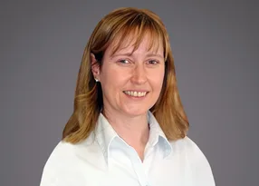 A smiling person with shoulder-length light brown hair, wearing a white shirt, facing the camera against a grey gradient background.