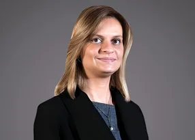 Portrait of a person with shoulder-length blond hair, wearing a dark blazer over a blue top, smiling softly against a grey background.