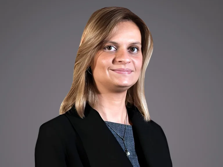 Person with shoulder-length blond hair, wearing a black blazer over a blue top, smiling against a neutral gray background.