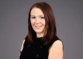 Portrait of a person with shoulder-length brown hair, wearing a black sleeveless top with metal rings, smiling against a neutral grey backdrop.