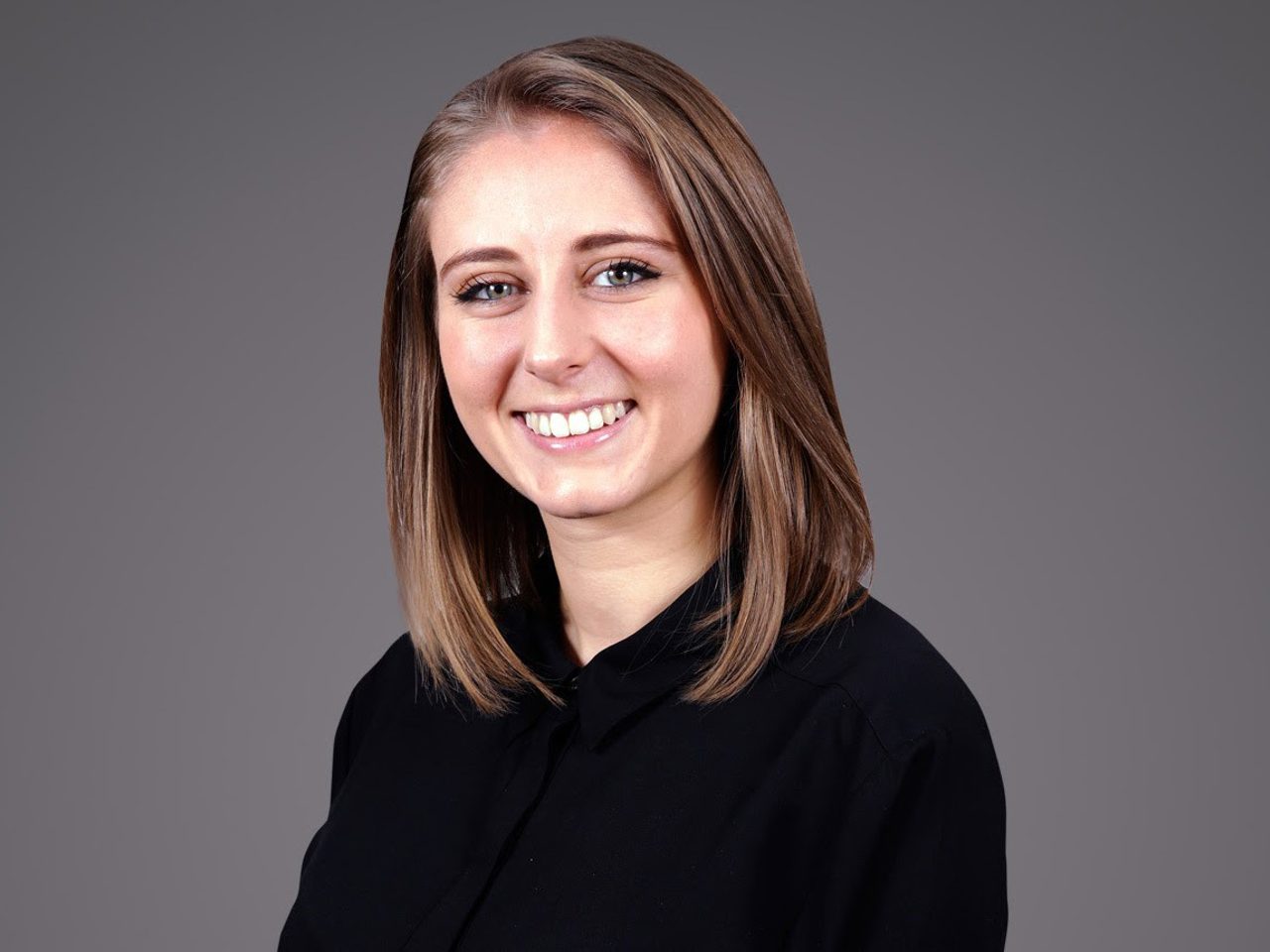 Portrait of a person with shoulder-length light brown hair, smiling, in a black top, against a neutral grey studio background.