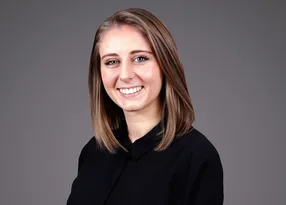 Portrait of a person with shoulder-length light brown hair, smiling, in a black top, against a neutral grey studio background.