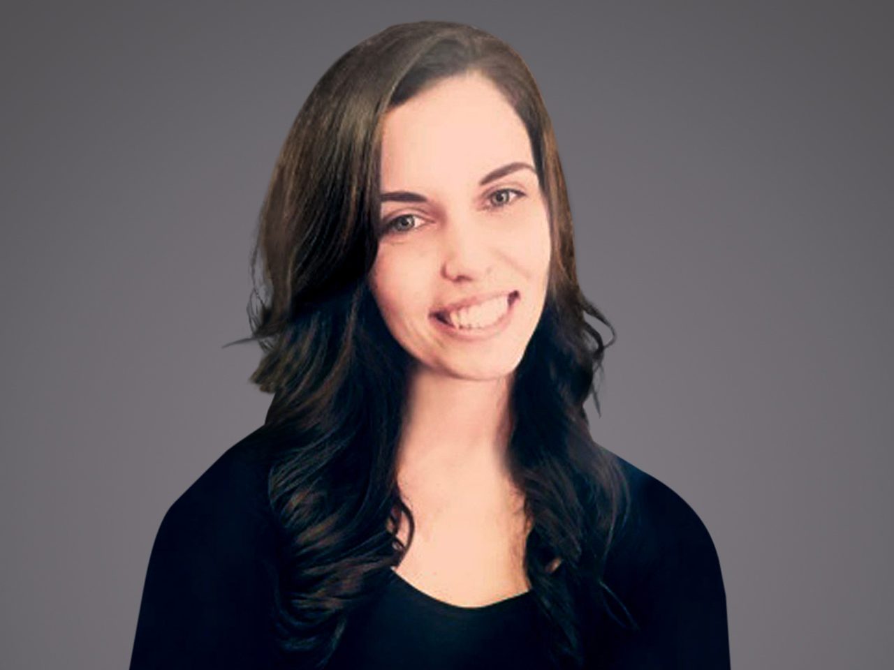 Portrait of a person with long, dark wavy hair wearing a black top, smiling with teeth showing, against a gray gradient background.