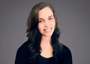 Portrait of a person with long dark hair, smiling, wearing a dark top, against a grey backdrop.
