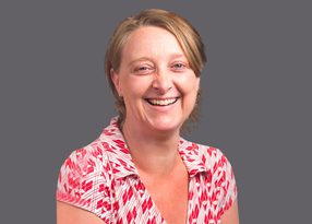 Smiling person with short blond hair wearing a red and white geometric patterned shirt, against a gray background.