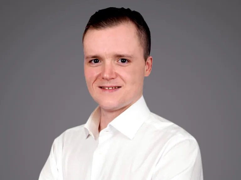 Porträt einer Person in weißem Hemd vor grauem Hintergrund; freundliches Lächeln, leicht seitlicher Blick.