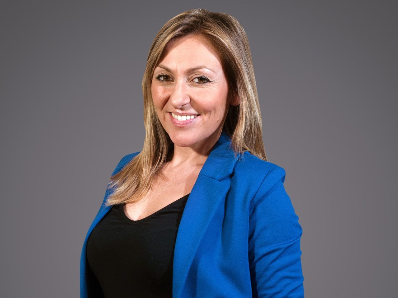 A smiling person with shoulder-length blonde hair wearing a blue blazer over a black top, set against a grey studio background.