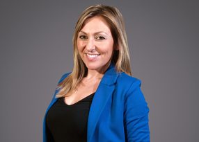 A smiling person with shoulder-length blonde hair wearing a blue blazer over a black top, set against a grey studio background.