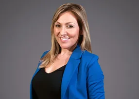 A smiling person with shoulder-length blonde hair wearing a blue blazer over a black top, set against a grey studio background.