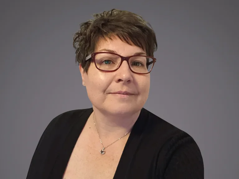 Porträt einer Person mit kurzen braunen Haaren, roter Brille, schwarzer Oberbekleidung und silberner Kette vor grauem Hintergrund, Blick leicht zur Seite.