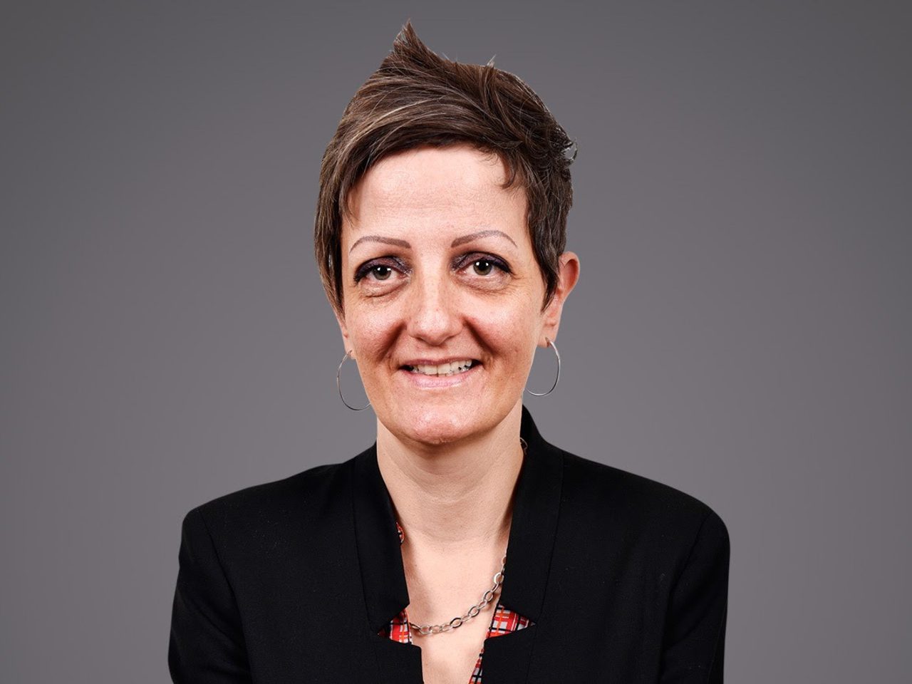 Portrait of a person with short hair, wearing a black blazer, hoop earrings and a silver chain necklace, smiling against a grey backdrop.