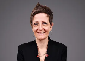Portrait of a person with short hair, wearing a black blazer, hoop earrings and a silver chain necklace, smiling against a grey backdrop.