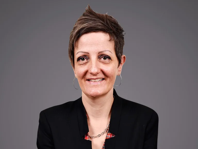 Portrait of a person with short hair, wearing a black blazer, hoop earrings and a silver chain necklace, smiling against a grey backdrop.