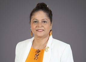Portrait of a person with dark hair in a bun, wearing orange top, white blazer, orange earrings, and a gold brooch, smiling against a gray backdrop.