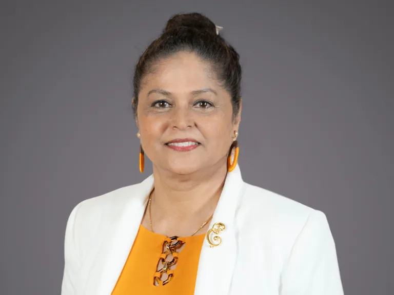 Portrait of a person with dark hair in a bun, wearing orange top, white blazer, orange earrings, and a gold brooch, smiling against a gray backdrop.