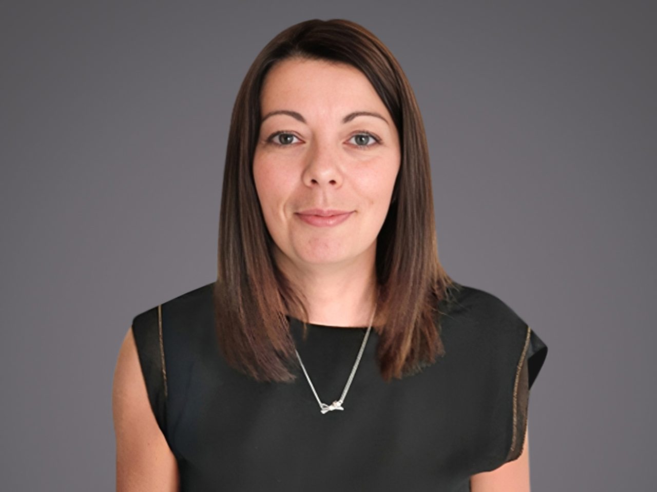 Persona con capelli castani a taglio medio, indossa top nero e una collana con ciondolo, sfondo grigio uniforme, guarda verso la fotocamera con leggero sorriso.