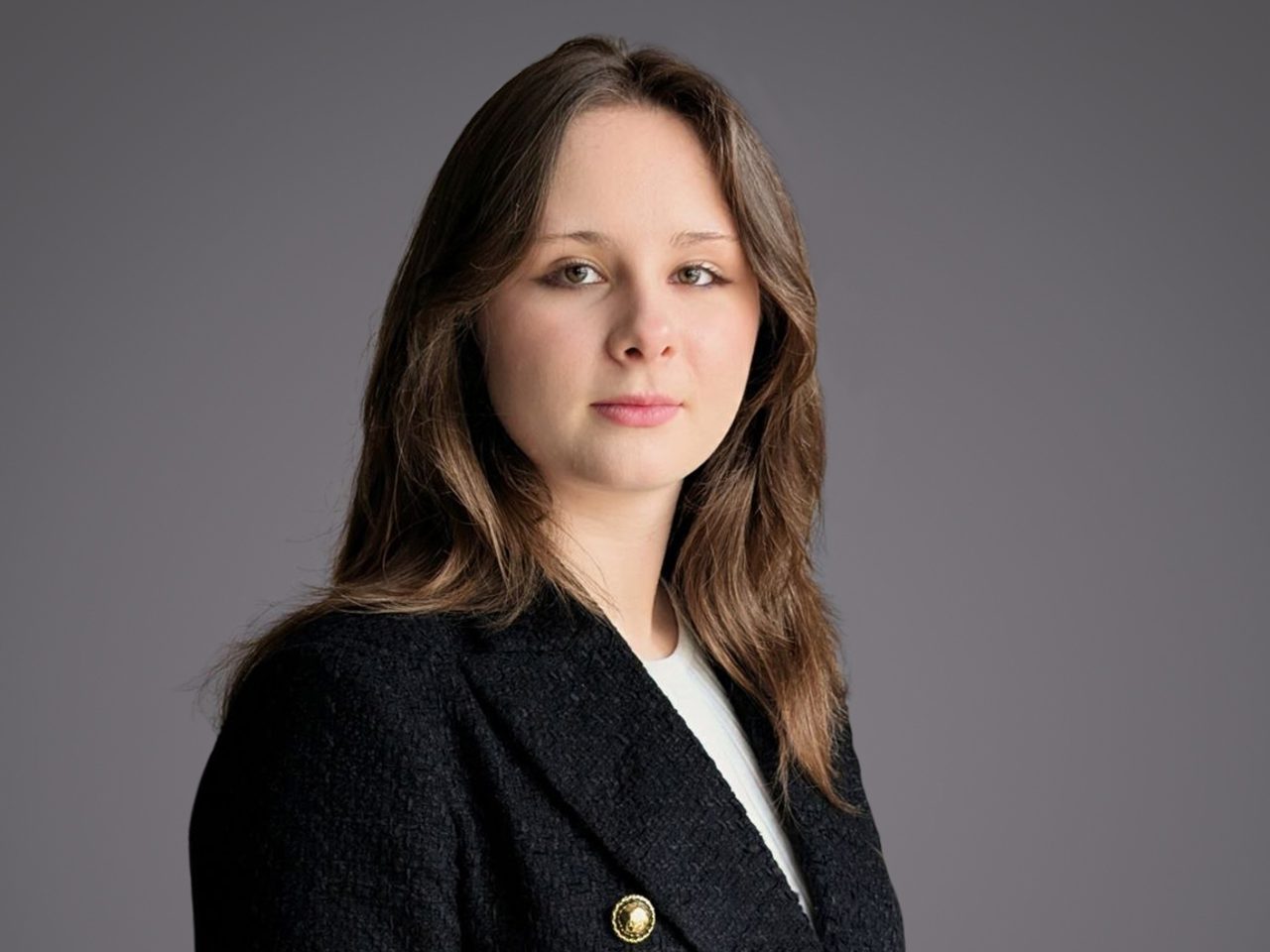 Portrait of a person with shoulder-length brown hair, wearing a dark blazer with a gold pin over a white top, posed against a neutral gray backdrop.