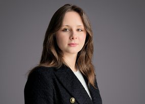 Portrait of a person with shoulder-length brown hair, wearing a dark blazer with a gold pin over a white top, posed against a neutral gray backdrop.