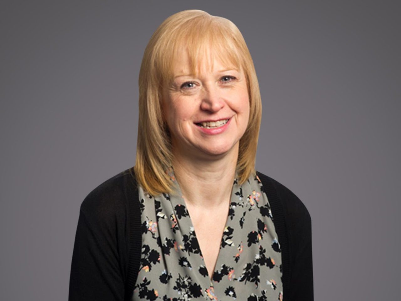 Smiling person with shoulder-length blonde hair, wearing a floral gray blouse and black cardigan, against a neutral gray studio background.