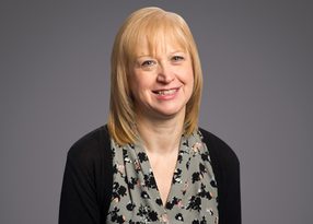 Smiling person with shoulder-length blonde hair, wearing a floral gray blouse and black cardigan, against a neutral gray studio background.