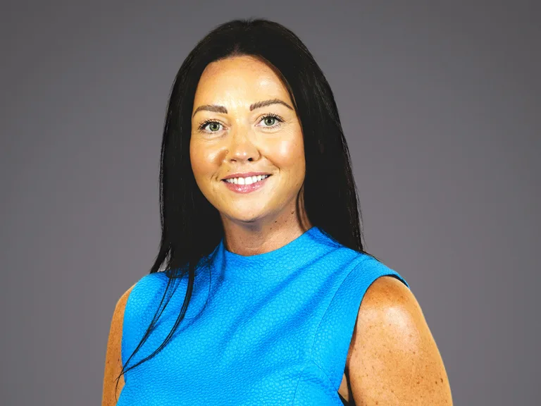 Femme souriante aux cheveux noirs, portant une robe bleue sans manches, posant contre un fond gris neutre.