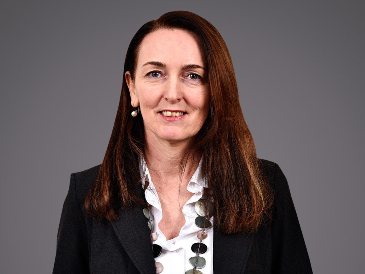 Personne portant veste noire et chemise blanche à volants, collier de disques et boucles d’oreilles, cheveux longs bruns, sourire, fond gris.