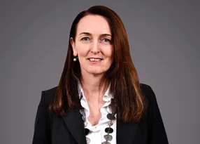 Portrait of a person with long brown hair, wearing a dark blazer over a white blouse and a circular disc necklace, smiling against a grey backdrop.