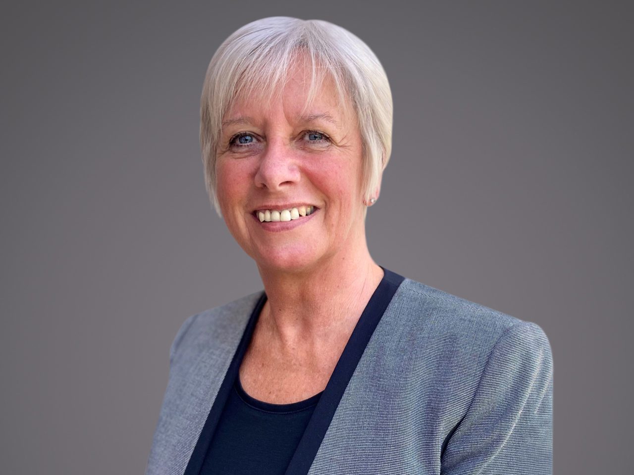 Smiling person with short gray hair, wearing a gray blazer over a navy top, against a neutral gray backdrop.