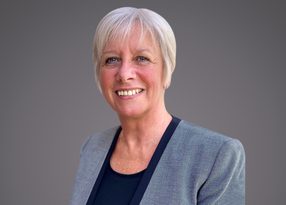 Smiling person with short gray hair, wearing a gray blazer over a navy top, against a neutral gray backdrop.