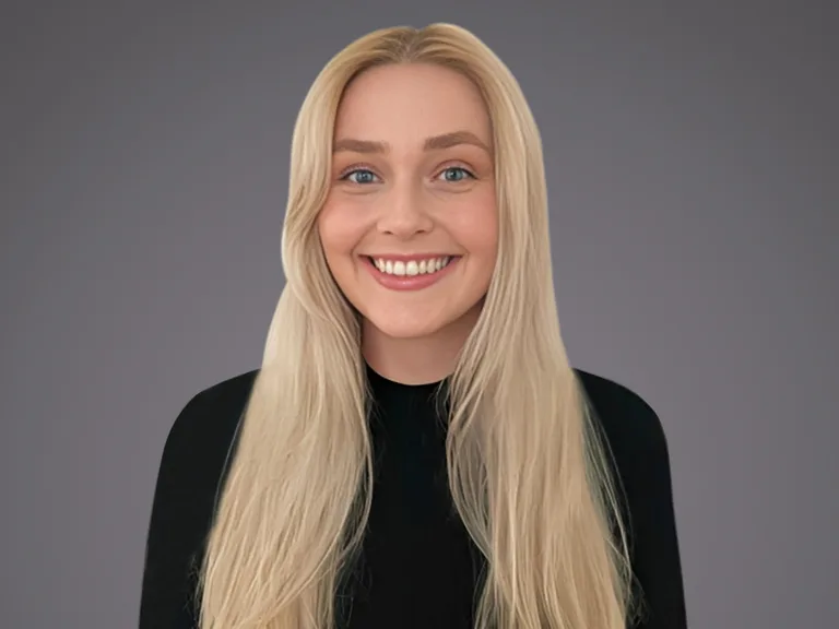 A person with long, straight blonde hair and blue eyes, wearing a black top, smiling at the camera against a plain grey background.