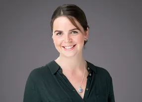 Smiling person with a dark green button-down blouse, short brown hair pulled back, wearing a silver necklace with a round iridescent pendant, against a gray studio backdrop.