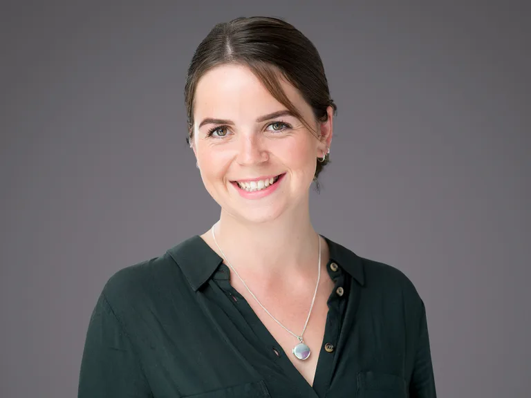 Porträt einer Person mit dunkelgrünem Hemd, braunen kurzen Haaren, freundlichem Lächeln; silberne Kette mit rundem Anhänger vor grauem Hintergrund.