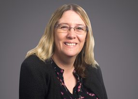 Portrait of a person with shoulder-length blond hair and glasses, smiling, wearing a black cardigan over a floral black blouse, against a gray gradient background.