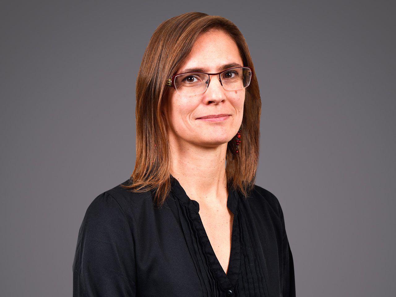 Personne debout, cheveux châtains mi-longs, portant des lunettes et une chemise noire, sourire discret, fond gris neutre.