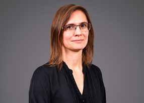 Personne debout, cheveux châtains mi-longs, portant des lunettes et une chemise noire, sourire discret, fond gris neutre.