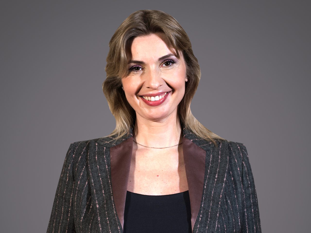 Smiling person with shoulder-length light brown hair, wearing a dark pinstripe blazer with satin lapels over a black top, against a grey backdrop.