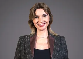 Smiling person with shoulder-length light brown hair, wearing a dark pinstripe blazer with satin lapels over a black top, against a grey backdrop.
