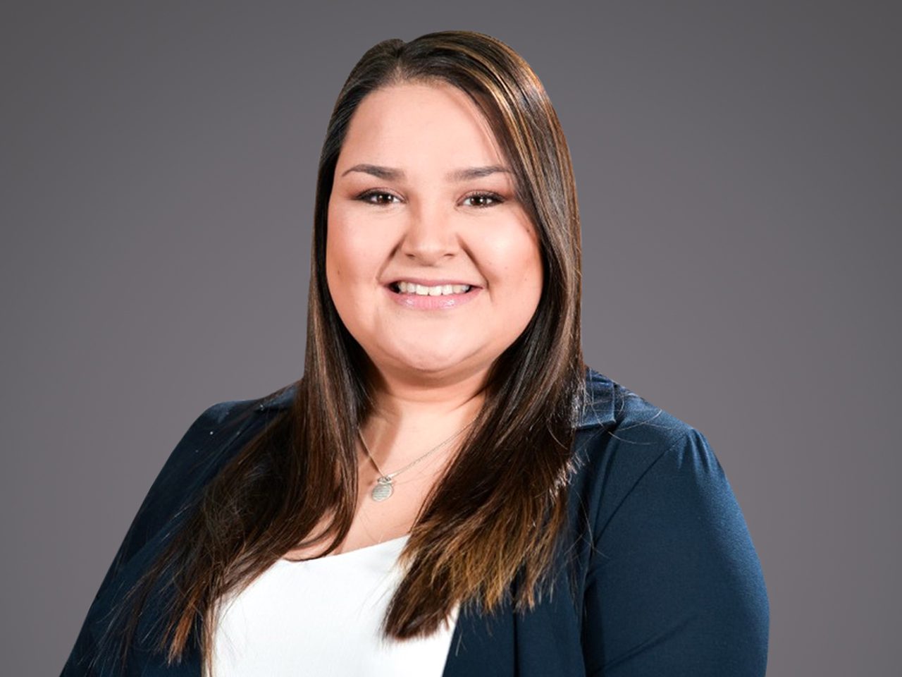 Persona sorridente con capelli lunghi castano scuri, indossa giacca blu scuro e top bianco, collana con ciondolo, sfondo grigio uniforme.