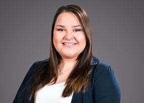 Smiling person with long dark hair wearing a navy blazer over a white top and a circular pendant, photographed against a gray studio background.