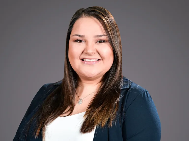 Smiling person with long dark hair wearing a navy blazer over a white top and a circular pendant, photographed against a gray studio background.