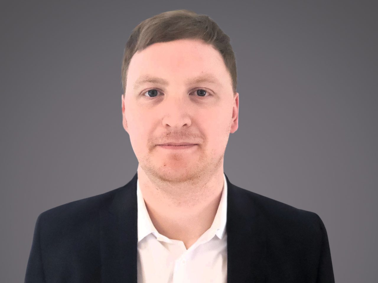 Portrait of a person with short light brown hair, wearing a white shirt and dark blazer, facing the camera against a grey gradient background.