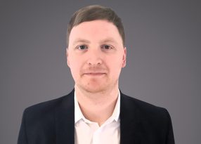 Portrait of a person with short light brown hair, wearing a white shirt and dark blazer, facing the camera against a grey gradient background.