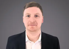 Portrait of a person with short light brown hair, wearing a white shirt and dark blazer, facing the camera against a grey gradient background.