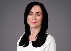 Portrait of a person with shoulder-length black hair and light skin, wearing a white blouse with a black rounded collar, against a gray background.