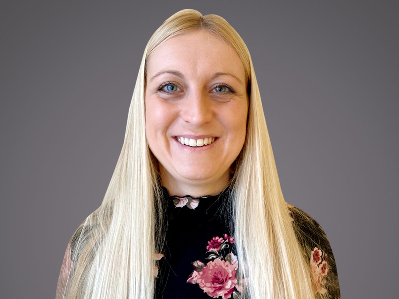 Portrait of a person with long straight blonde hair, smiling, wearing a black floral top, against a neutral gray background.