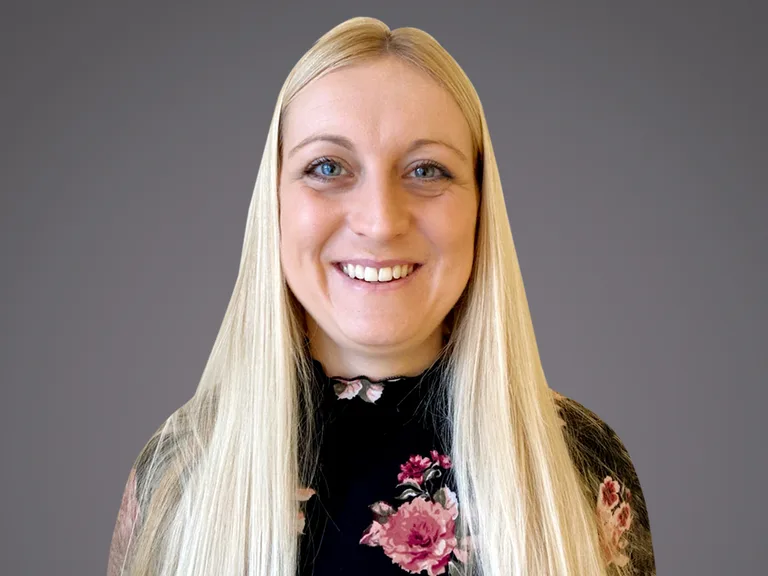 Portrait of a person with long straight blonde hair, smiling, wearing a black floral top, against a neutral gray background.