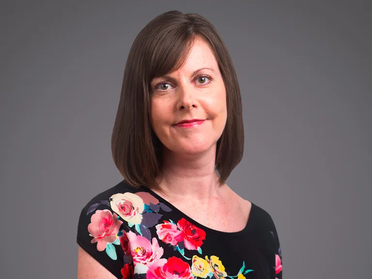 Person with shoulder-length brown hair, wearing a black top with a colorful floral print, posed against a gray studio background.