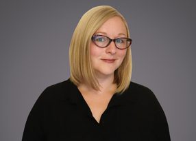 Headshot of a person with shoulder-length blond hair, red-framed glasses, and a black collared top, smiling softly against a gray gradient background.