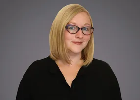 A person with shoulder-length blonde hair and red-framed glasses, wearing a black top, against a grey gradient background.