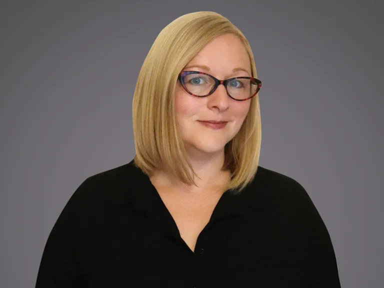 Headshot of a person with shoulder-length blond hair, red-framed glasses, and a black collared top, smiling softly against a gray gradient background.